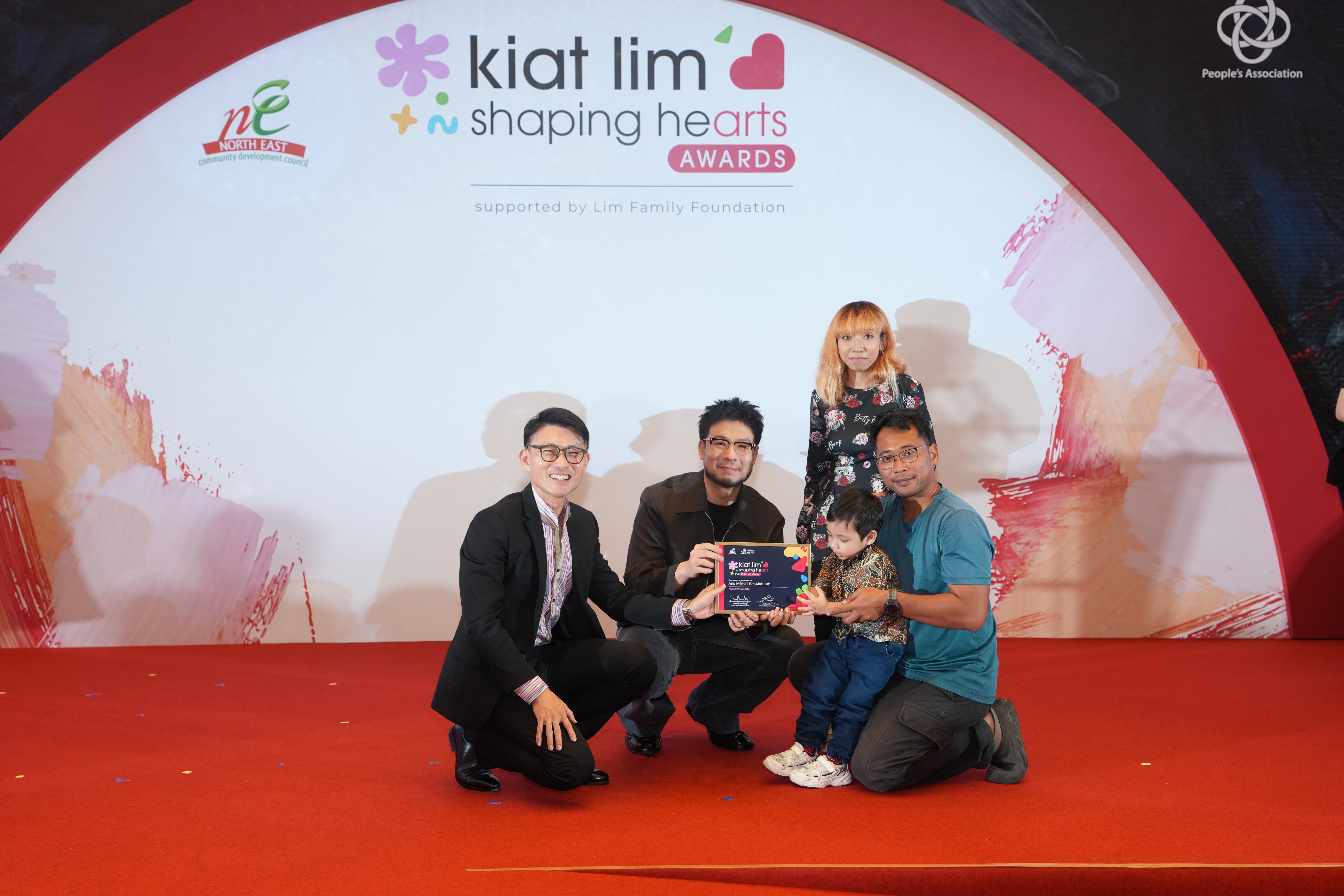 Award recipient standing on stage with the presenter, holding their certificate and posing for a group photo during the award ceremony, with a backdrop reading ‘Kiat Lim-Shaping Hearts Awards'.
